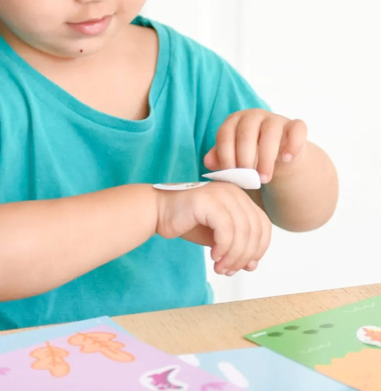 A young child applies a round sticker from the Poppik Sticker Cards Set - Baby – Dinosaurs to their forearm while sitting at a table, enjoying colorful dinosaur-themed sticker sheets during fun preschool activities.