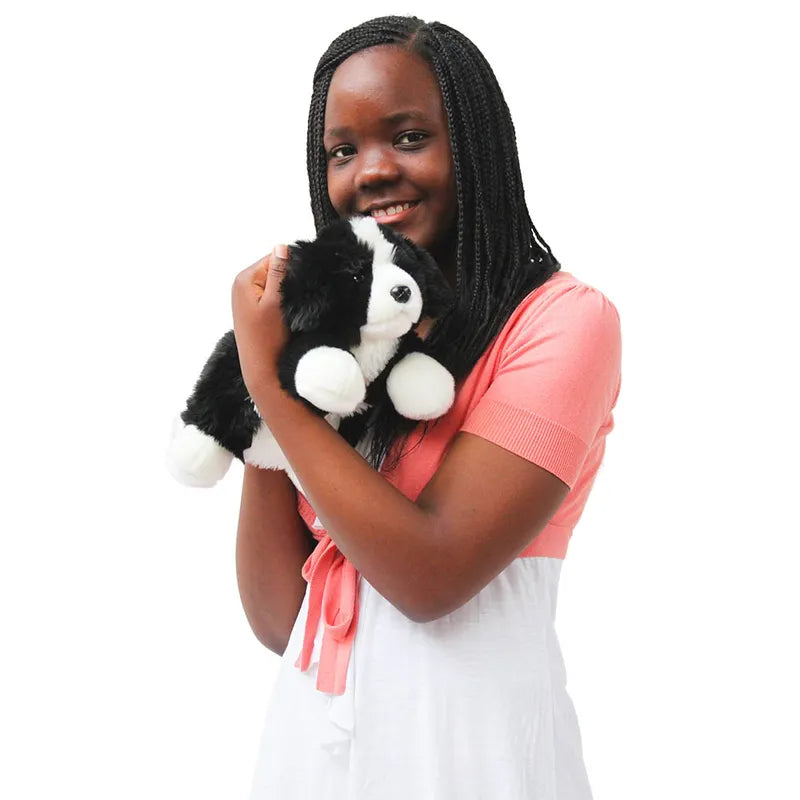 A smiling person with braided hair, in a pink short-sleeved top over a white dress, holds The Puppet Company Full-bodied Hand Puppet Border Collie against a white background.