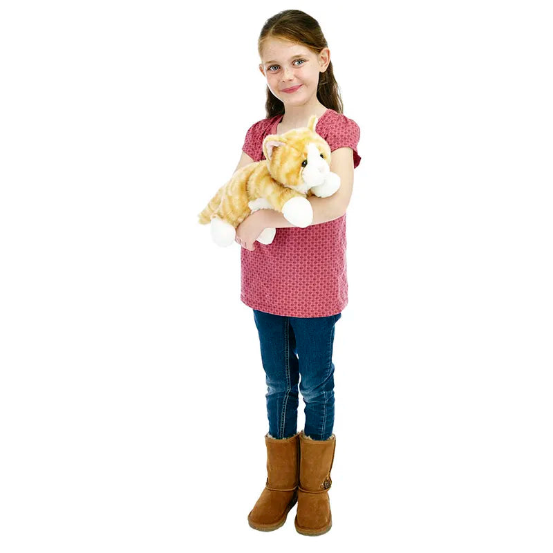 A young girl in a red patterned shirt, blue jeans, and brown boots is holding her cuddly The Puppet Company Full-bodied Hand Puppet Cat Ginger. Her hair is tied back, and she smiles at the camera.