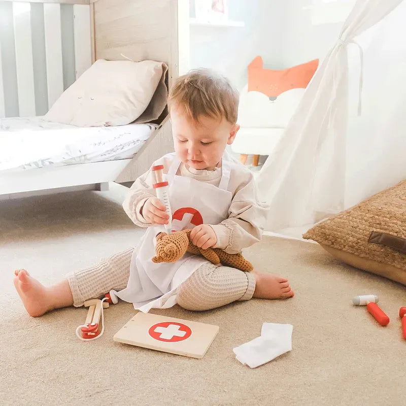 A toddler sits on a carpeted floor, using the Bigjigs Doctor's Kit to examine a stuffed animal, while wearing a white apron with a red cross. The room features warm colors, a bed, and a canvas play tent.