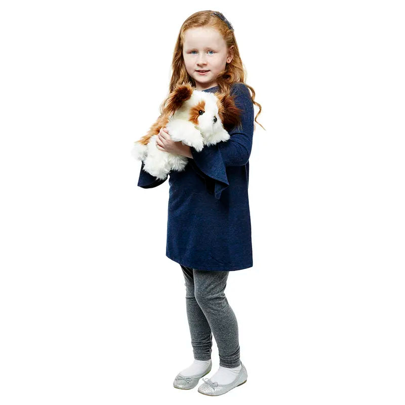 A young girl with long red hair, in a navy blue dress, gray leggings, and silver shoes, stands holding The Puppet Company Full-bodied Hand Puppet Dog against a white background.