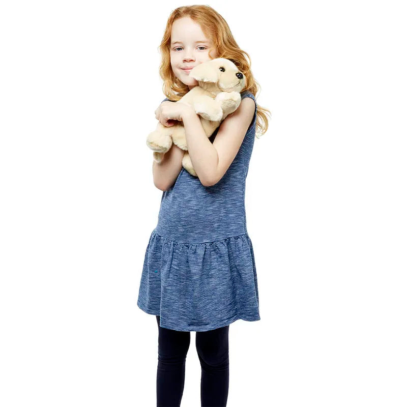 A young girl with long red hair, clad in a blue dress and black leggings, smiles as she hugs The Puppet Company Full-bodied Hand Puppet Labrador, standing against a white background.