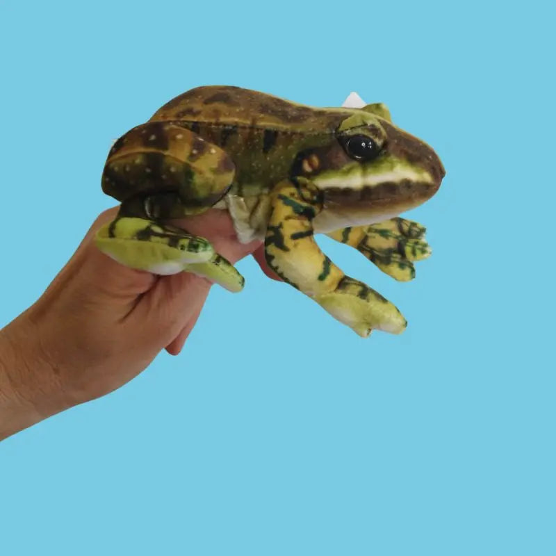 A hand holds The Puppet Company Green Frog Finger Puppet from the Little Creatures Range against a plain light blue background, ideal for storytime fun.
