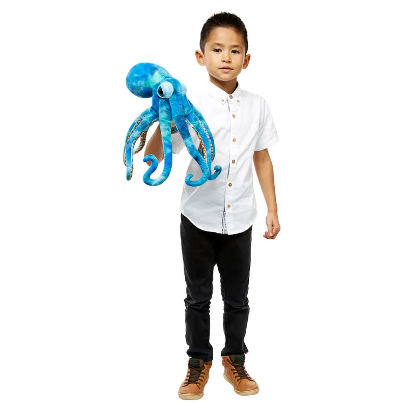 A young boy, dressed in a white button-up shirt, dark pants, and brown shoes, stands against a plain white background holding The Puppet Company Large Creature Octopus—a perfect companion for imaginative play. He gazes forward with a neutral expression.