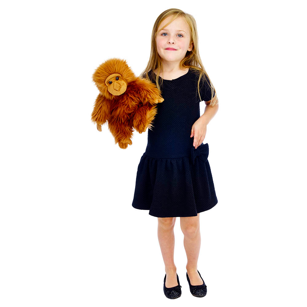 A young girl in a black dress and shoes holds The Puppet Company Full-bodied Hand Puppet Orangutan, standing against a white background.