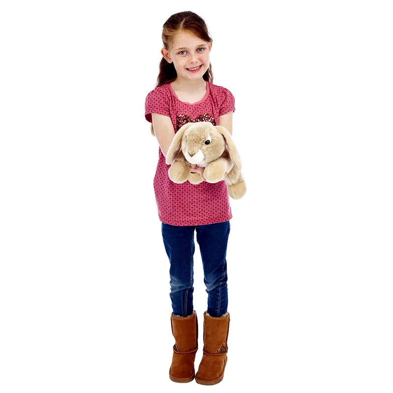 A young girl in a pink shirt, blue jeans, and brown boots smiles while holding The Puppet Company Full-bodied Hand Puppet Rabbit with floppy ears. The background is white.