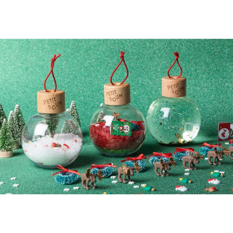 Christmas Sensory Baubles—three clear ornaments with wooden tops and red strings, filled with fake snow, red beads, and green liquid—are displayed on a green surface among mini trees, reindeer, and festive décor.