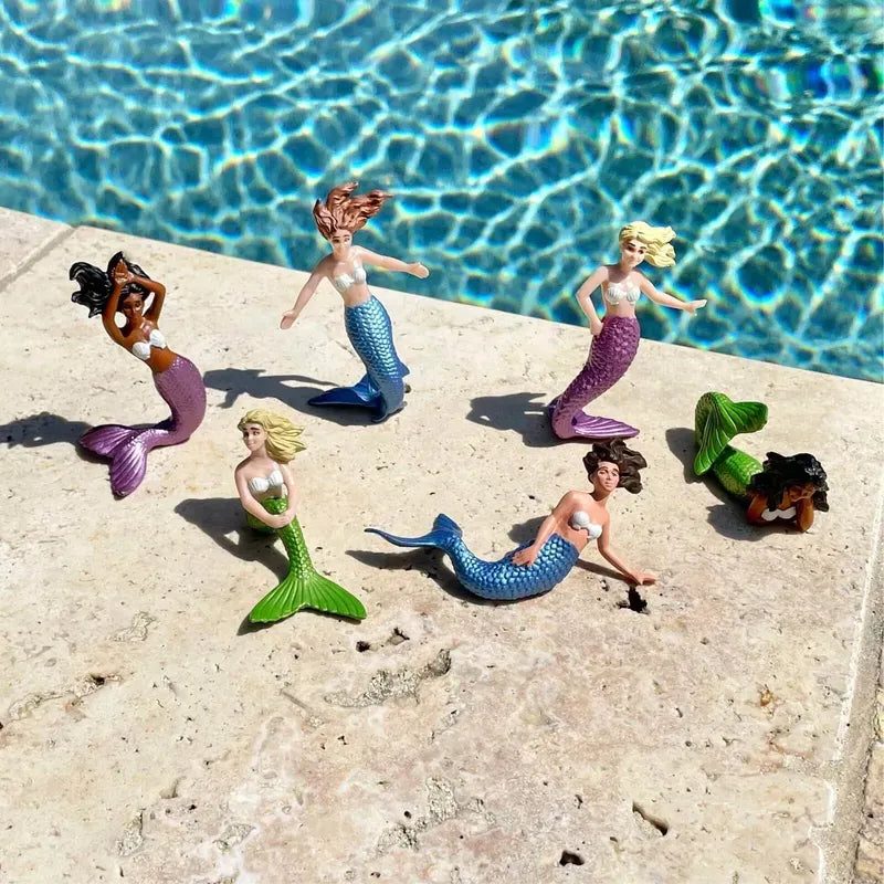 Five diverse TOOB® Figurines Mermaids with colorful tails are positioned on a concrete edge near a sparkling pool.