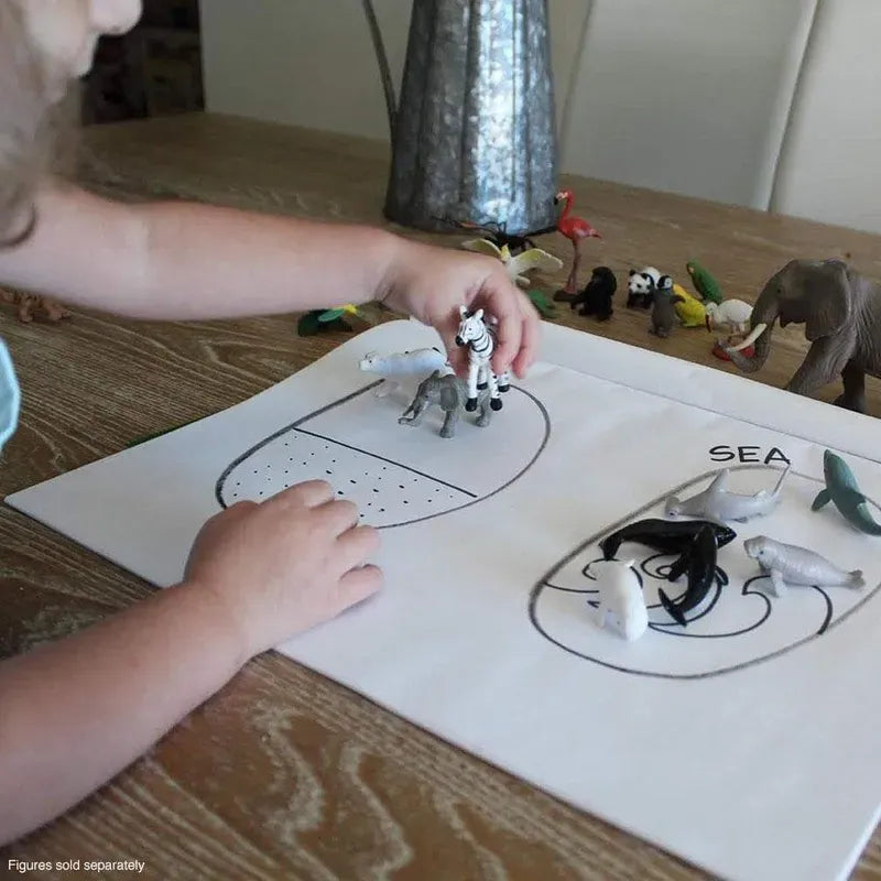 A child's hands placing TOOB® Figurines Baby Sea Life on a labeled activity sheet stating "sea," with various other TOOB® figurines around on a wooden table.