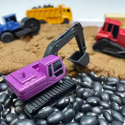 TOOB® Figurines Construction Vehicles construction scene with a purple excavator on black beans intended to simulate rocky terrain, surrounded by a red bulldozer, a blue truck, and yellow dump truck figurines on sandy ground.