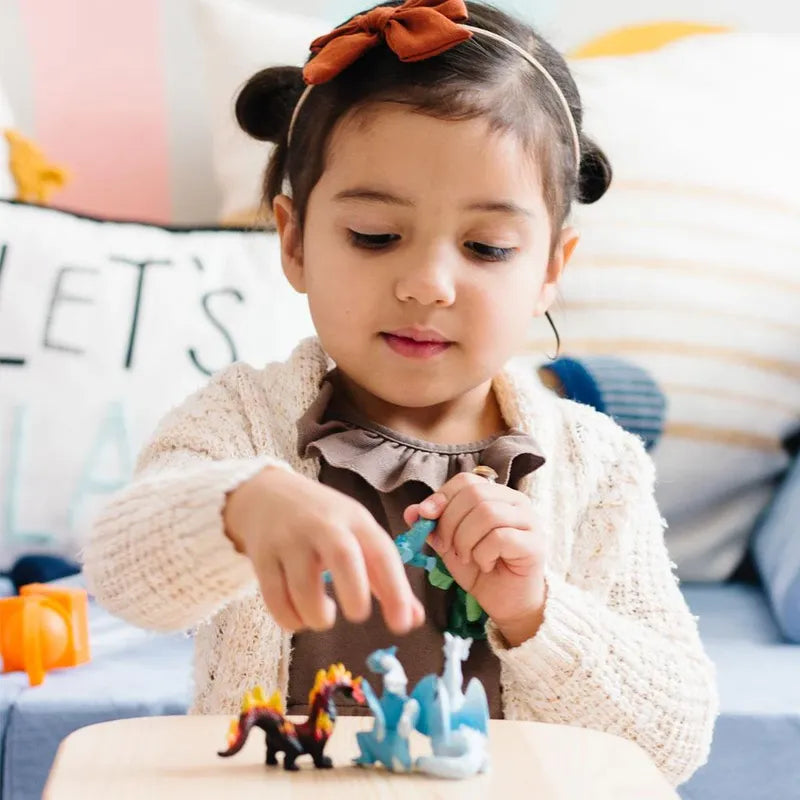 A young girl with a headband plays with TOOB® Figurines Dragons of the Elements on a table, surrounded by colorful pillows and a plush toy.