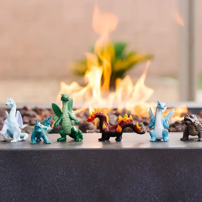 A collection of colorful TOOB® Figurines Dragons of the Elements arranged in a row on a surface, with a blurred fire pit in the background, creating a dramatic backdrop.