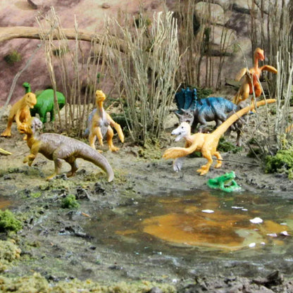 A detailed TOOB® Figurines Feathered Dinos diorama featuring various hand-painted dinosaur figurines, including a stegosaurus and velociraptor, set in a realistic prehistoric scene with a small pond, rocks.