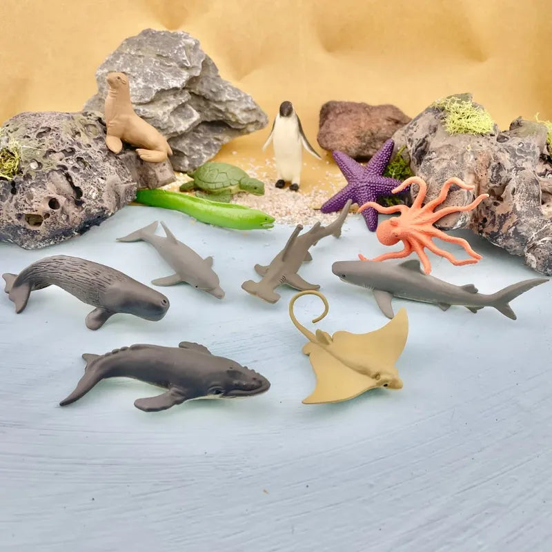 A collection of TOOB® Figurines Ocean including dolphins, sharks, starfish, a penguin, and a seal, displayed among rocks and foliage on a light blue surface.