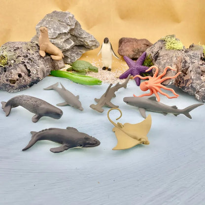 A collection of TOOB® Figurines Ocean including dolphins, sharks, starfish, a penguin, and a seal, displayed among rocks and foliage on a light blue surface.