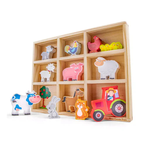 A wooden toy set featuring a storage box with compartments holding colorful farm animal figures, a tree, a red tractor, and a farmer. Some animal figures are placed in front of the box.