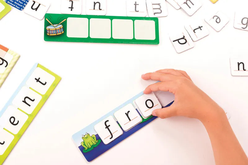 A child arranges letter tiles to spell "frog" on a card featuring a cartoon frog from the Orchard Toys Match And Spell set, with additional tiles and cards scattered on a white surface.