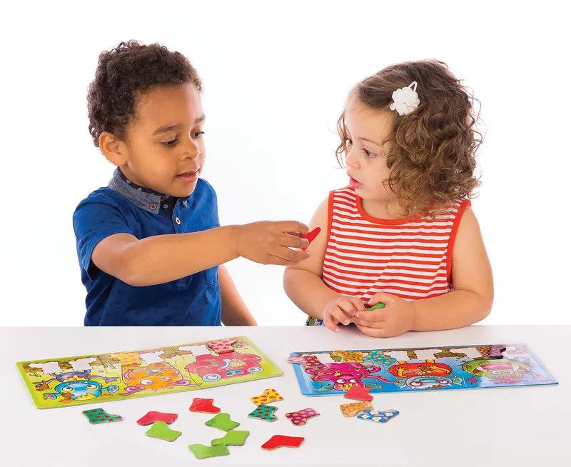 Two young children sit at a table, engaging with vibrant Orchard Toys Smelly Wellies jigsaw puzzles. The boy, dressed in a blue shirt, passes a puzzle piece to the girl in a red and white striped dress. The puzzles showcase colorful animal designs.