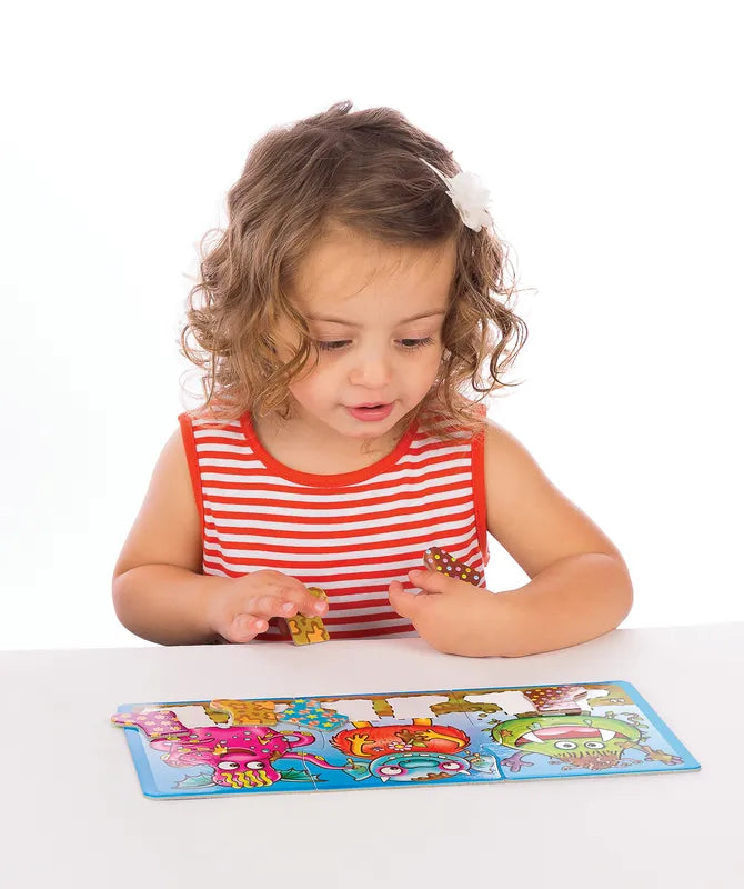 A young child with curly hair and a white hair clip is wearing a red and white striped shirt. She is sitting at a table and playing with the colorful Orchard Toys Smelly Wellies puzzle, which features cartoon animals. The background is plain white.