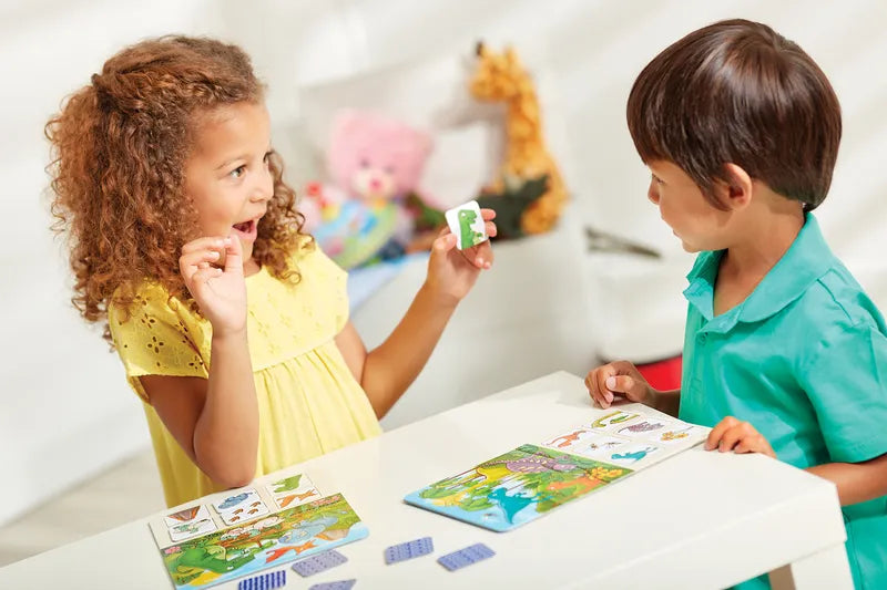 Two children sitting at a table are playing Orchard Toys Dinosaur Lotto, using cards and a game board. The girl, wearing a yellow dress, is excitedly holding a card, while the boy in a teal shirt watches with interest. In the background, there are various toys and a stuffed giraffe.