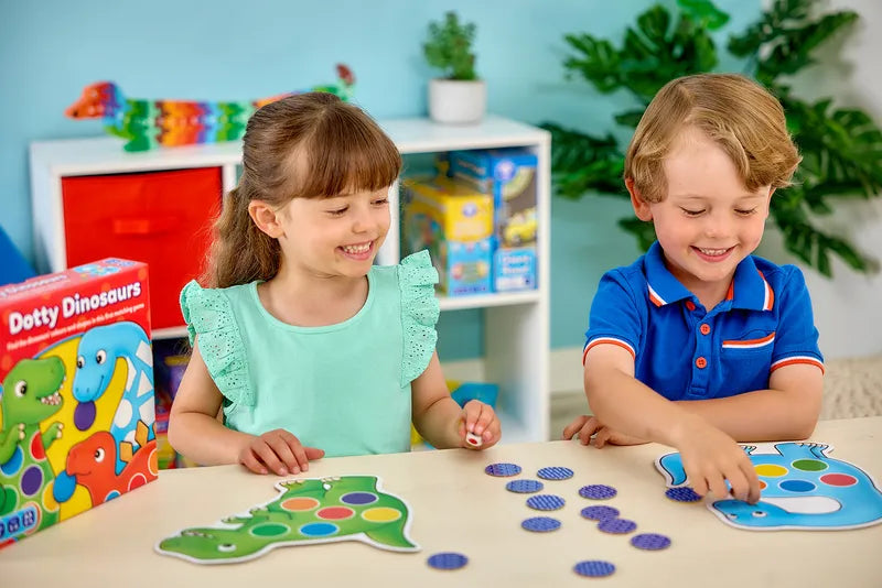 Two smiling children are enjoying a board game featuring dinosaur-shaped cards and blue tokens. The box, labeled "Orchard Toys Dotty Dinosaurs," sits on the table. In the background, there are plants and colorful shelves filled with more games.