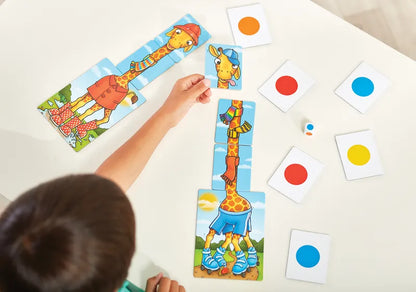 A child assembling the Orchard Toys Giraffes In Scarves puzzle on a white table. The puzzle pieces depict cartoon giraffes wearing outfits, and nearby are cards with colored circles and a multi-colored die.