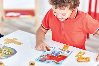 A young child with curly hair, wearing a red polo shirt, is playing Orchard Toys Magic Maths. He is placing a small card onto the board, which has colorful illustrations. Other game pieces and cards are scattered on the table.