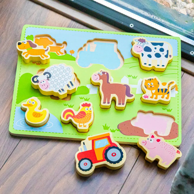 The New Classic Toys Chunky Farm Puzzle is a colorful wooden puzzle that features various farm animals and a red tractor. This delightful wooden toy includes pieces such as a dog, sheep, cow, duck, rooster, horse, cat, pig. Some pieces are placed in their corresponding slots on a green board, while others are outside.