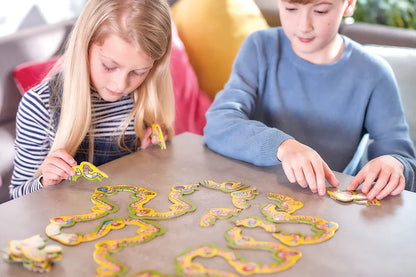 Two children are sitting at a table assembling the vibrant Orchard Toys Wiggly Words puzzle, featuring winding paths. The girl, wearing a striped shirt, and the boy, in a blue shirt, are deeply focused on fitting the pieces together. Bright cushions add a cheerful touch to the background.