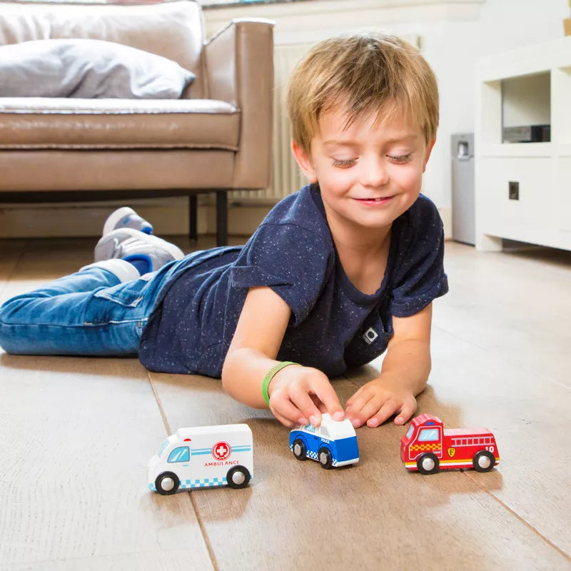 A young boy, one of the many little car lovers, is lying on the floor of a living room, smiling and playing with toddler toys. The New Classic Toys Vehicles set includes a white ambulance, a blue police car, and a red fire truck. The room has a sofa and a white TV cabinet in the background.