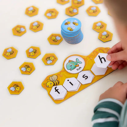 A child's hands arrange hexagonal tiles from the Orchard Toys Buzz Words set on a board. The board shows an image of a fish with the letters "f", "i", "s", "h" forming the word "fish," and bee-themed tiles are scattered around.