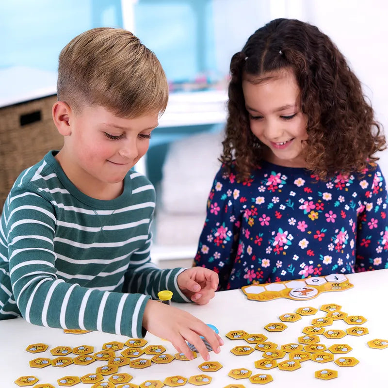 Two children, a boy and a girl, are sitting at a table playing Orchard Toys Buzz Words with its distinctive yellow hexagonal tiles. The boy, dressed in a striped shirt, is concentrating on the game while the girl, wearing a floral dress, smiles as she observes.