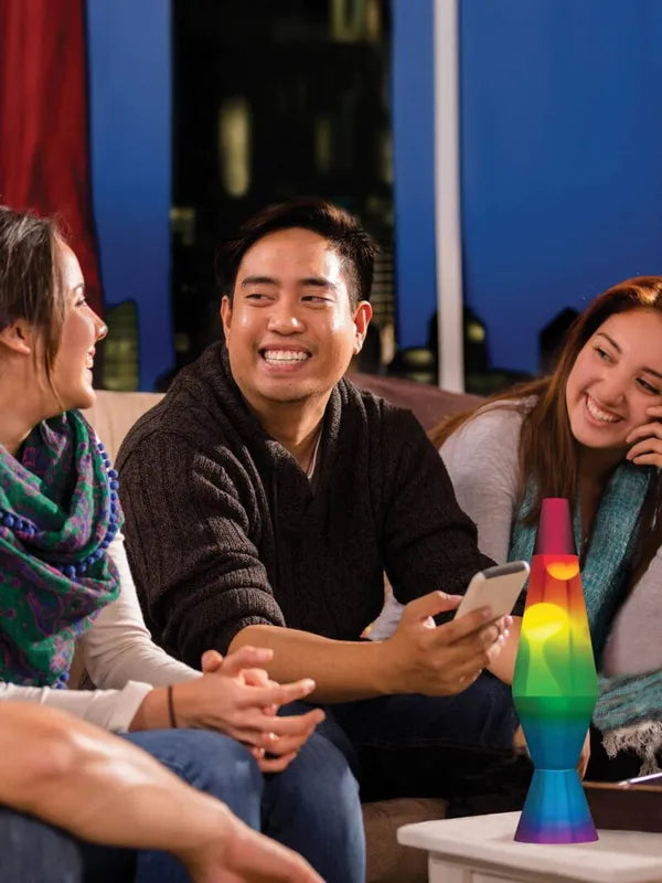 Three people sit on a couch, smiling and chatting. One holds a smartphone, while a 14.5" Rainbow LAVA Lamp on the table adds vibrant colors as a sensory accessory. The city lights through the window create a cozy urban atmosphere in the background.