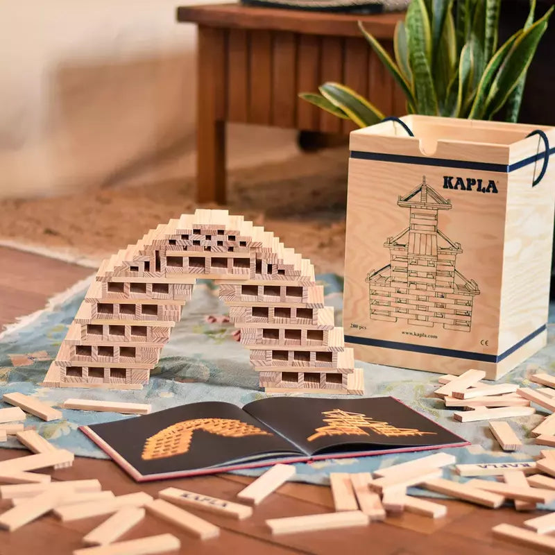 An intricately assembled wooden block arch structure, created using pieces from the KAPLA® Construction 280 Planks in Wooden Box, displayed on a floor with additional blocks scattered around and design booklets in the foreground, all showcasing.