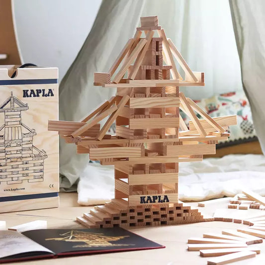 The Intricately stacked wooden blocks from a KAPLA® Construction 280 Planks in Wooden Box, forming a complex, multi-layered tower structure reminiscent of architectural marvels, with the guidebook and a few unused pieces.