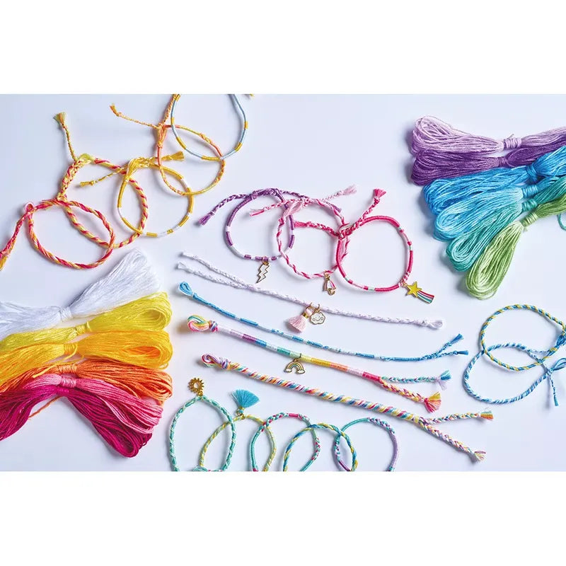 Displayed on a white surface, the Janod 30 Rainbow Wrap Bracelets To Make kit showcases a collection of colorful friendship bracelets and charms. The bracelets feature various designs and rainbow colors, including twisted and braided styles. Additionally, the kit includes bundles of embroidery floss in different shades, making it an irresistible creative set for crafting enthusiasts.