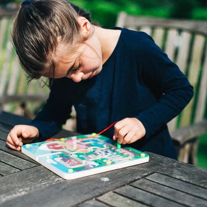 A girl is playing with the Bigjigs Farmyard Maze Puzzle on a wooden table, honing her fine motor skills as she navigates through a farm maze.