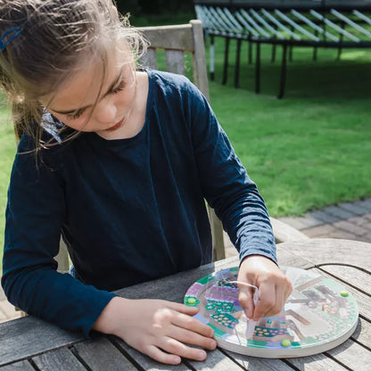 A young girl is playing with the Bigjigs Flower Garden Maze Puzzle on a wooden table, enhancing her fine motor skills.