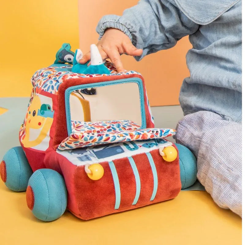 Amidst an orange backdrop, a child in light blue clothing enjoys the vibrant Lilliputiens I'm Discovering Activities Car Safari plush toy, featuring vehicle shapes, patterns, a friendly face, and a tiny side mirror — an ideal choice for a baby's first toy.