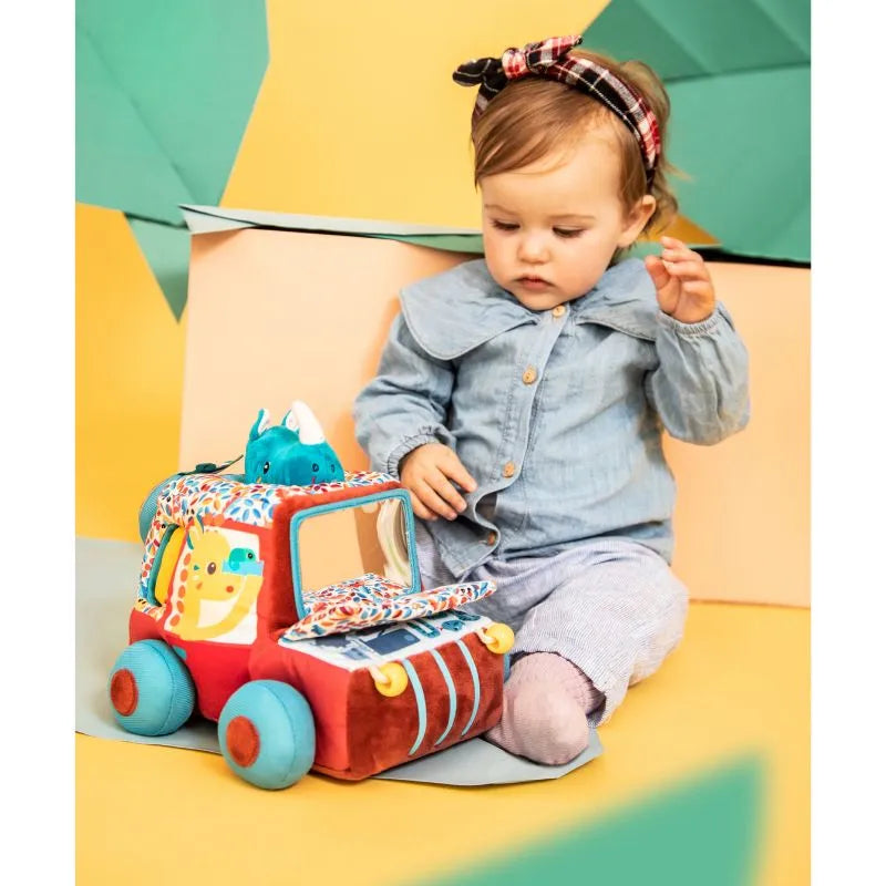 A toddler in a denim shirt and headband sits on a colorful background, playing with the Lilliputiens I'm Discovering Activities Car Safari. This toy, adorned with vibrant patterns and a small dinosaur figure, is perfect for capturing the joy of discovering baby's first gift.