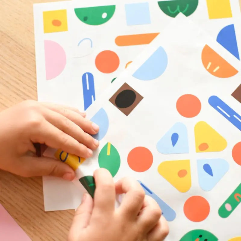 A child peels vibrant animal stickers from the Poppik Sticker Cards Set - Baby – Animals on a wooden table, with extra colorful sheets underneath—ideal for fun preschool activities.