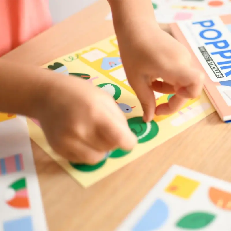 A child places colorful animal stickers from the Poppik Sticker Cards Set - Baby – Animals onto a yellow sheet at a wooden table, with portable sticker cards and the set nearby—perfect for fun preschool activities.