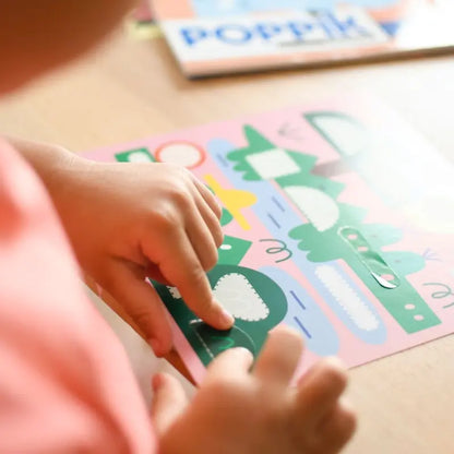 A child is making an art project at a table, using the Poppik Sticker Cards Set - Baby – Animals to peel and stick green and white shapes onto a colorful sheet—ideal for preschool crafts or on-the-go animal-themed sticker fun.