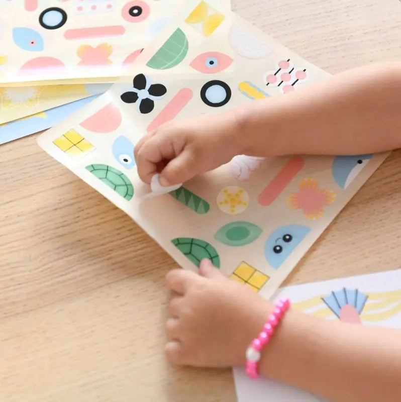 A child peels stickers from the Poppik Sticker Card Set Baby – Ocean on a wooden table, wearing a pink beaded bracelet and selecting colorful ocean-themed shapes that help preschoolers develop fine motor skills.