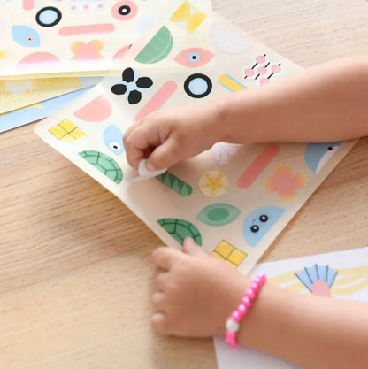 A child peels stickers from the Poppik Sticker Card Set Baby – Ocean on a wooden table, wearing a pink beaded bracelet and selecting colorful ocean-themed shapes that help preschoolers develop fine motor skills.