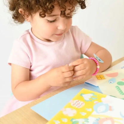 A young child with curly hair, in a light pink shirt and beaded bracelet, enjoys the Poppik Sticker Card Set Baby – Ocean at a table with colorful papers and stickers, developing fine motor skills through engaging sticker activities.
