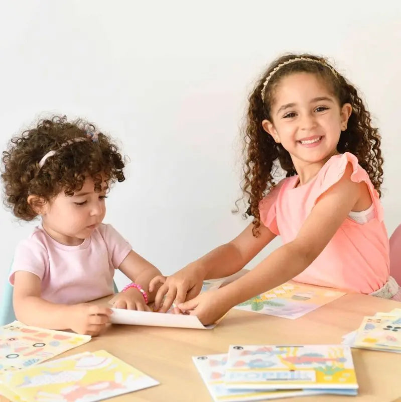 Two preschool children with curly hair in light pink clothes and headbands sit at a table with colorful papers. One girl smiles at the camera, while the other enjoys the Poppik Sticker Card Set Baby – Ocean to develop fine motor skills.