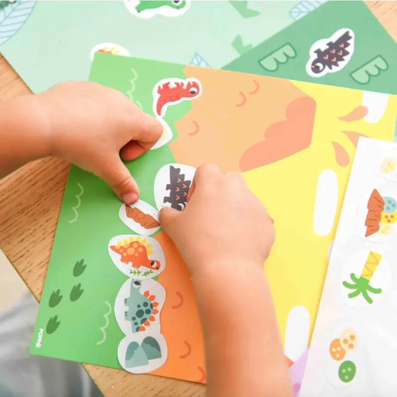 A child’s hands place colorful stickers from the Poppik Sticker Cards Set - Baby – Dinosaurs onto an activity sheet at a wooden table, with additional portable sticker cards featuring playful dinosaur designs visible—ideal for creative preschool fun.