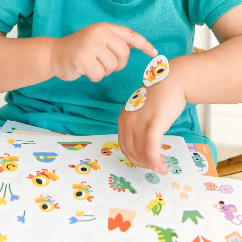A child in a teal shirt decorates their arm with chick and other baby animal stickers from the Poppik Sticker Cards Set - Baby - Baby Animals, perfect for preschool activities. The playful sticker sheet is portable and fun.