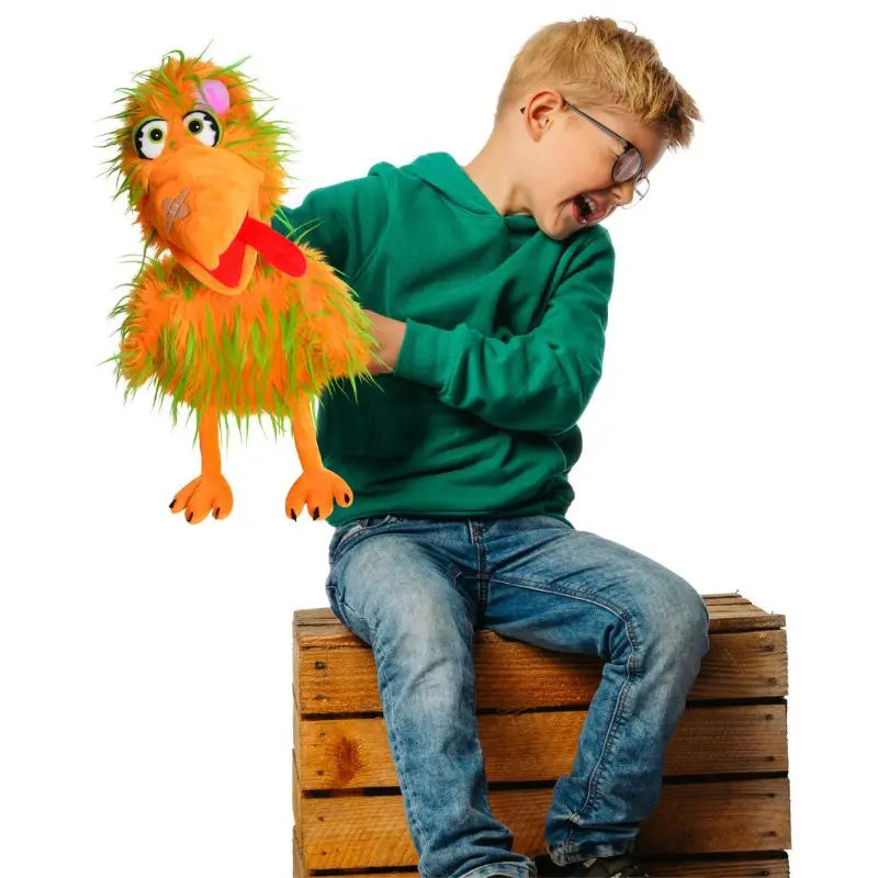 A young boy in glasses and a green hoodie sits on a wooden box, playfully holding the Living Puppets Bernie the Bird Hand Puppet, a colorful puppet with an orange beak and green-yellow feathers, ready for storytime fun.